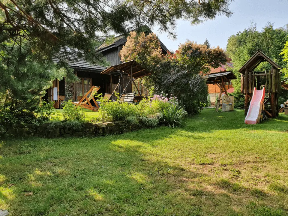 Plastikowa różowa zjeżdżalnia oraz dwie huśtawki położone w ogrodzie, pośród nich możemy znaleźć różnego rodzaju kwiaty