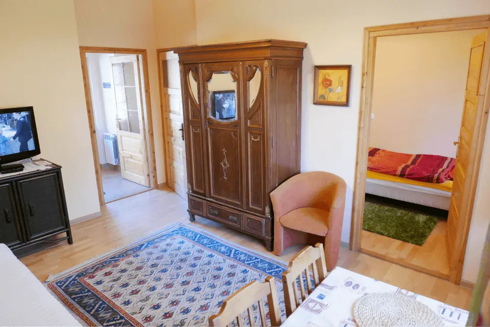Apartament pod jodełką, widok na cały apartament, znajdziemy tutaj salon z dużą szafą, telewizorem oraz wejściami do poszczególnych pokoi, nie brakuje również dywanu i stolika z dwoma krzesłami